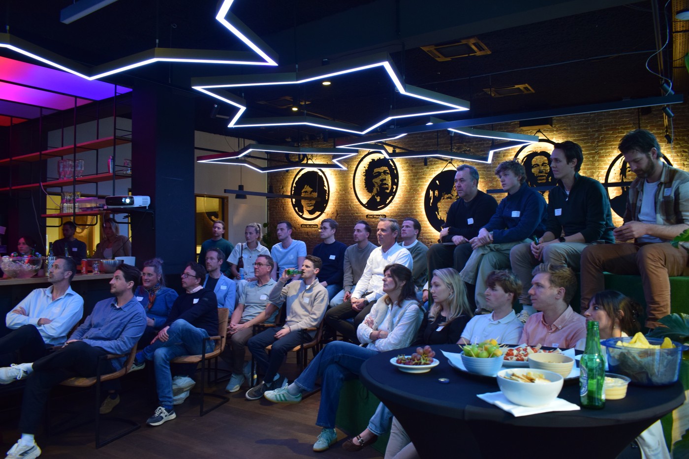 Audience seated in a modern event space listening to a presentation, with neon ceiling lights and a relaxed networking setup